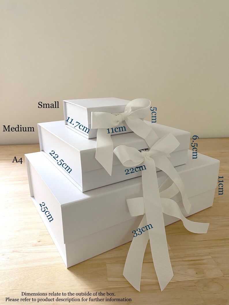 May include: Four white gift boxes of different sizes stacked on top of each other. The boxes are tied with white ribbon and labeled with their dimensions in centimeters. The boxes are on a light brown wooden surface.