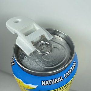 Puede incluir: Un abridor de latas de plástico blanco fijado en la parte superior de una lata azul y amarilla. La lata tiene el texto "CAFEÍNA NATURAL" impreso en ella.