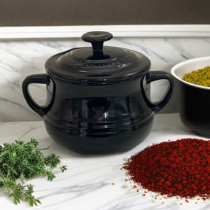 May include: A black ceramic cooking pot with a lid and two handles. The pot sits on a marble surface next to a pile of red spices, green herbs, and a bowl of green spices. The pot is a kitchenware item.