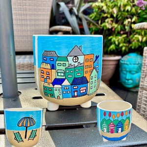 May include: Three ceramic plant pots with hand-painted designs. The largest pot features a cityscape with colourful houses against a blue sky. The smaller pots depict beach scenes with umbrellas, chairs, and beach huts. All pots have a beige base and white feet.