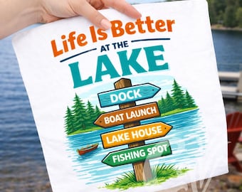 May include: White square towel with the text "Life Is Better at the Lake." The design includes a lake scene with a wooden signpost pointing to the dock, boat launch, lake house, and fishing spot. A small boat is on the water.