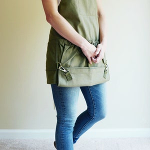 May include: A person wearing a green canvas apron with a white t-shirt and blue jeans. The apron has a large pocket and two smaller pockets on the sides. The apron has a brown leather strap and a metal buckle.