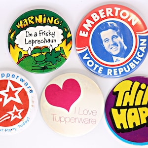 May include: Five round button pins with various designs. One pin is green with a cartoon monster and the text "WARNING: I'm a Frisky Leprechaun". Another pin is red, white, and blue with the text "EMBERTON VOTE REPUBLICAN" and a portrait of a man. A third pin is red with white stars and the text "Vote Tupperware Date Your Party Today!". A fourth pin is white with a pink heart and the text "I Love Tupperware". The last pin is purple with a yellow text that says "THINK HAPPY".
