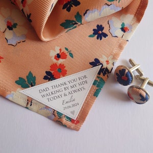 Peut inclure: Un mouchoir de poche de couleur pêche avec un motif floral et une étiquette blanche qui dit "DAD, THANK YOU FOR WALKING BY MY SIDE TODAY & ALWAYS. Emilia 29.06.2024". Le mouchoir de poche est associé à un jeu de boutons de manchette avec un motif floral similaire.