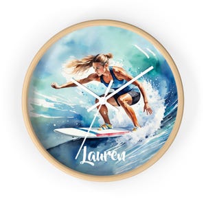 May include: Round wall clock with a watercolor illustration of a surfer riding a wave. The surfer is wearing a blue top and black shorts. The clock has a light wood frame and white clock hands. The name "Lauren" is written on the bottom.
