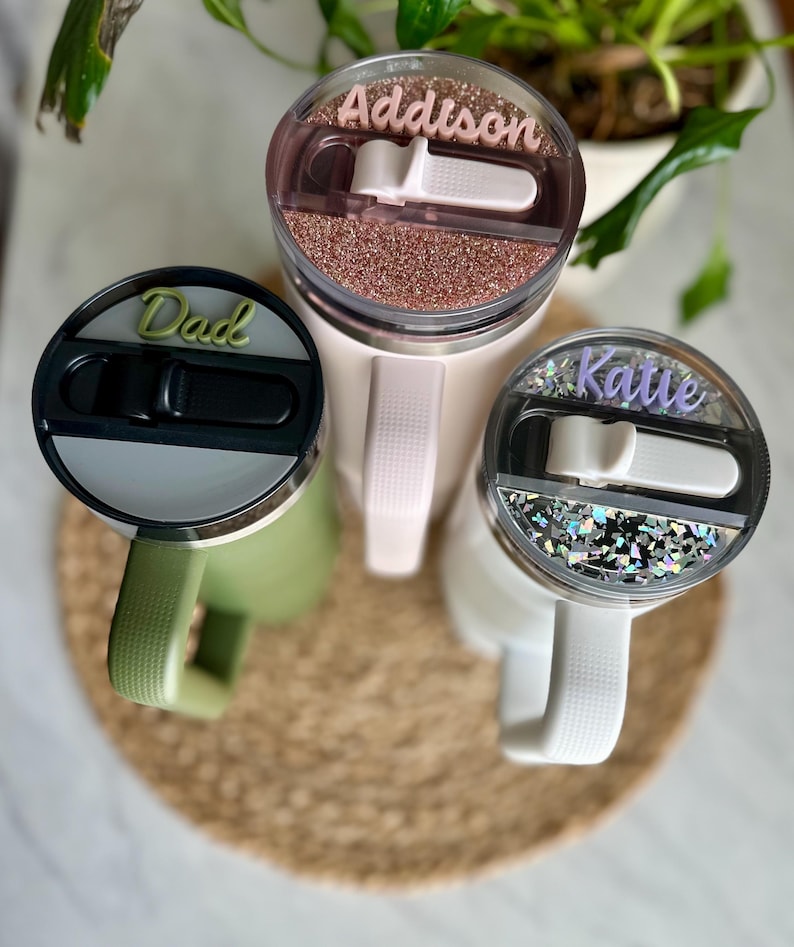 May include: Three personalized stainless steel tumblers with lids. The tumblers are green, pink, and white. The lids have glitter and the names "Dad", "Addison", and "Katie" written on them.