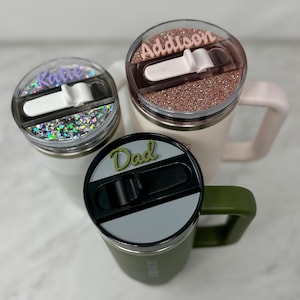 May include: Three personalized stainless steel travel mugs with different colored lids and names on them: Katie, Addison, and Dad. The mugs have a slider lid with a silicone seal.