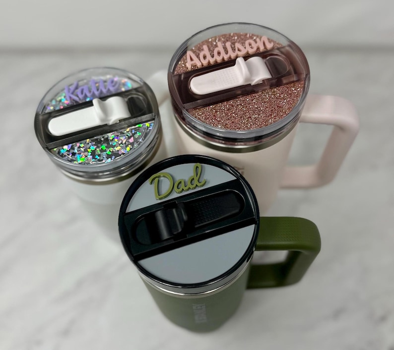 May include: Three personalized stainless steel travel mugs with different colored lids and names on them: Katie, Addison, and Dad. The mugs have a slider lid with a silicone seal.
