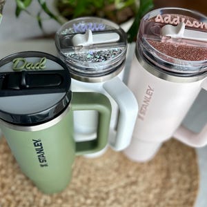 May include: Three stainless steel travel mugs with personalized lids. The mugs are green, white, and pink. The lids have the names "Dad", "Katie", and "Addison" written on them. Each mug has the Stanley logo on the side.