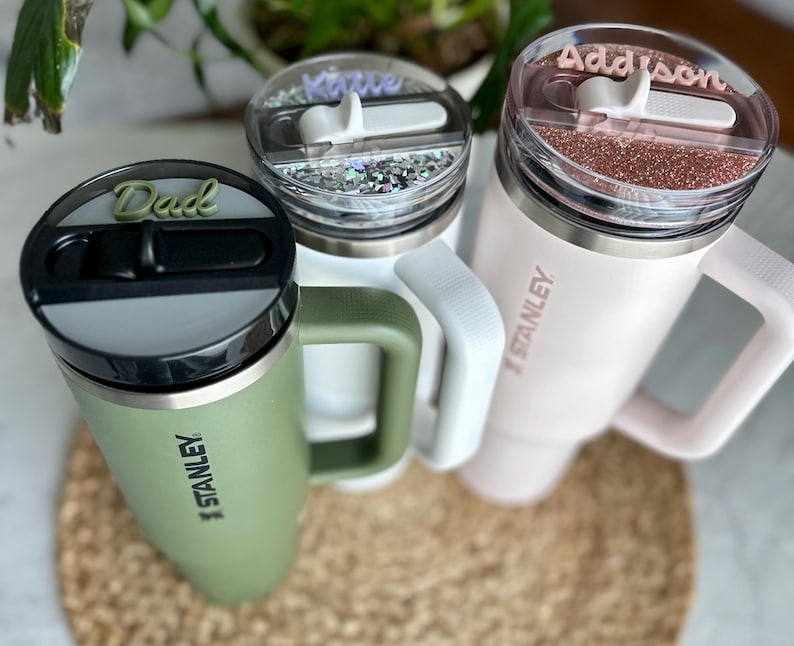 May include: Three stainless steel travel mugs with personalized lids. The mugs are green, white, and pink. The lids have the names "Dad", "Katie", and "Addison" written on them. Each mug has the Stanley logo on the side.