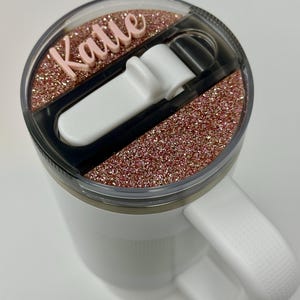 May include: White travel mug with a pink glitter lid and a black lid with a white slider. The lid has the name "Katie" written on it in pink.
