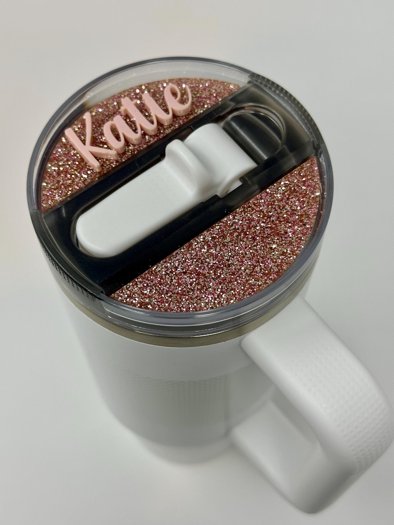 May include: White travel mug with a pink glitter lid and a black lid with a white slider. The lid has the name "Katie" written on it in pink.