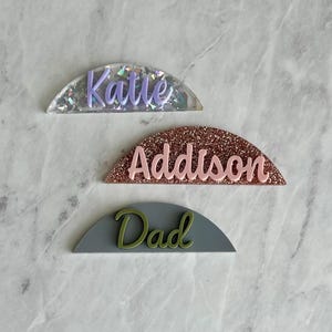May include: Three half-circle name tags with glitter backgrounds. The top tag is silver with purple glitter and the name "Katle" written in purple. The middle tag is rose gold with pink glitter and the name "Addison" written in pink. The bottom tag is gray with green glitter and the name "Dad" written in green.
