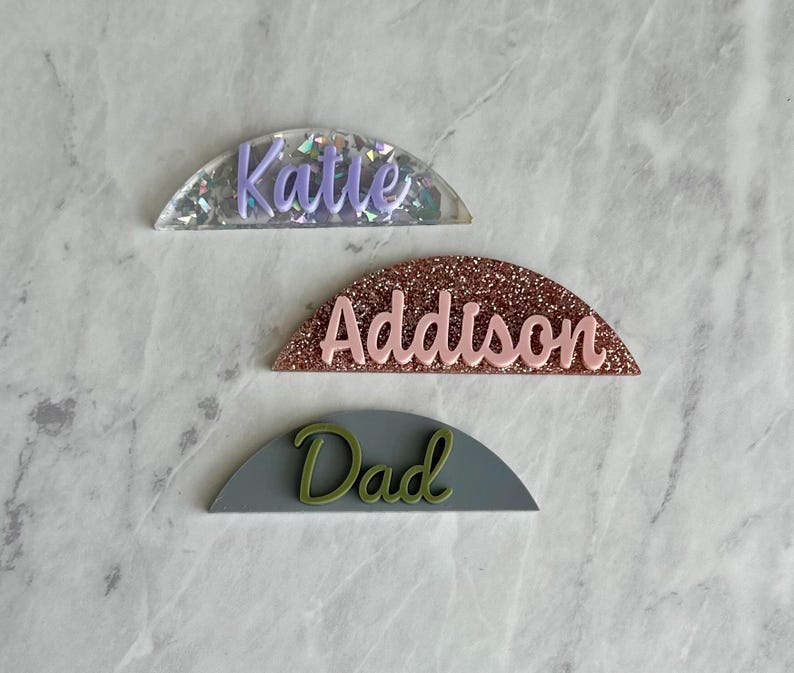 May include: Three half-circle name tags with glitter backgrounds. The top tag is silver with purple glitter and the name "Katle" written in purple. The middle tag is rose gold with pink glitter and the name "Addison" written in pink. The bottom tag is gray with green glitter and the name "Dad" written in green.