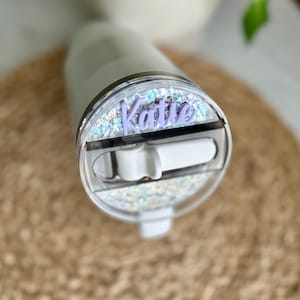 May include: A white tumbler with a clear lid featuring a silver glitter design and a purple name tag that reads "Katie".