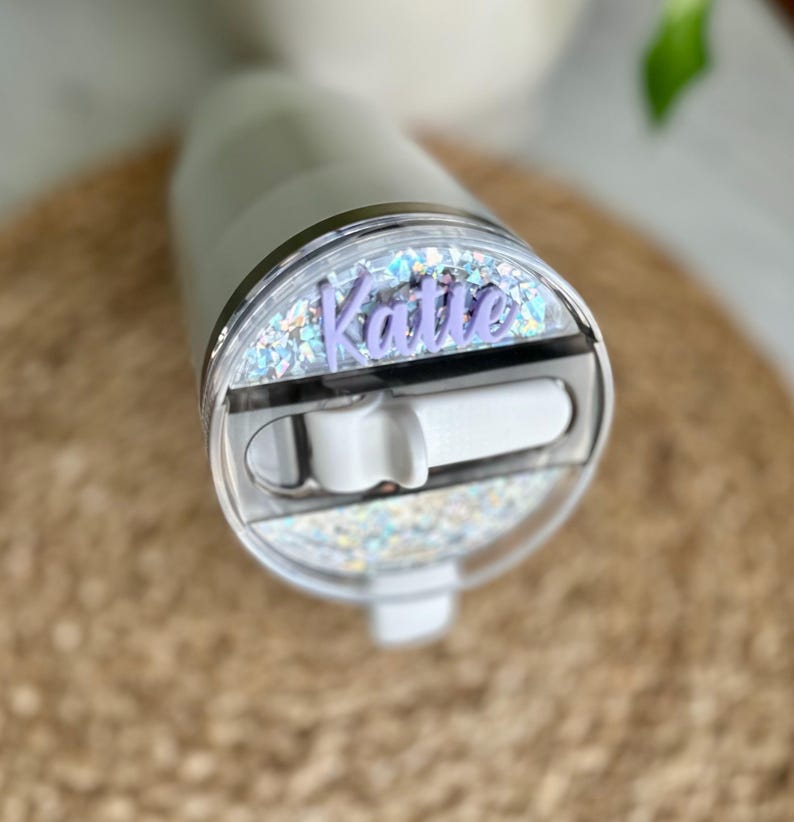 May include: A white tumbler with a clear lid featuring a silver glitter design and a purple name tag that reads "Katie".