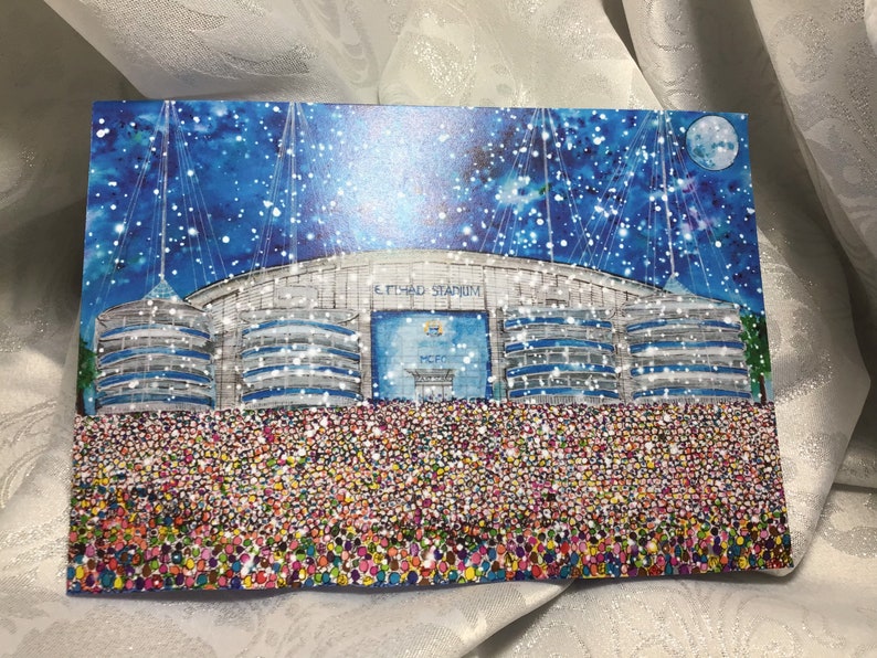 May include: A Christmas card featuring a snowy night scene of the Etihad Stadium in Manchester, England. The stadium is lit up and surrounded by a crowd of people. The card has a blue background with white snowflakes and a full moon.