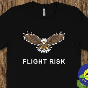May include: Black t-shirt with a white eagle graphic and the text "FLIGHT RISK" in white. A yellow circle with a black cat jumping over a blue ring and the text "CAT IN ORBIT" is in the lower right corner.