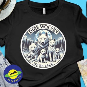 May include: Black t-shirt with a graphic of a pack of white dire wolves in a forest setting. The graphic includes the text "Dire Wolves We're Back".