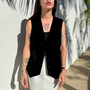 May include: A black velvet vest with a button-up front, paired with a white skirt. The vest has a V-neck design and is sleeveless. The outfit is worn outdoors in natural light.