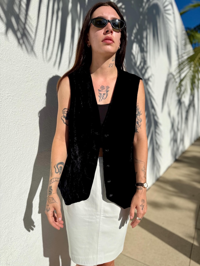 May include: A black velvet vest with a button-up front, paired with a white skirt. The vest has a V-neck design and is sleeveless. The outfit is worn outdoors in natural light.