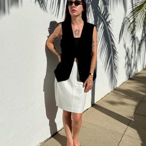 May include: A black sleeveless vest paired with a white skirt. The outfit is completed with black sunglasses and black flip-flops. The background features a white wall with palm tree shadows.