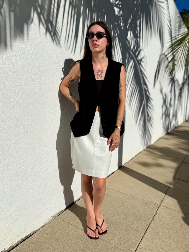 May include: A black sleeveless vest paired with a white skirt. The outfit is completed with black sunglasses and black flip-flops. The background features a white wall with palm tree shadows.