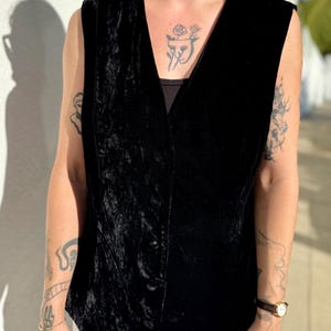 May include: A black velvet vest with a V-neck and button closure. The vest has a slightly textured appearance and is sleeveless. The garment is worn over a black top and white pants. The vest is a classic piece of apparel.