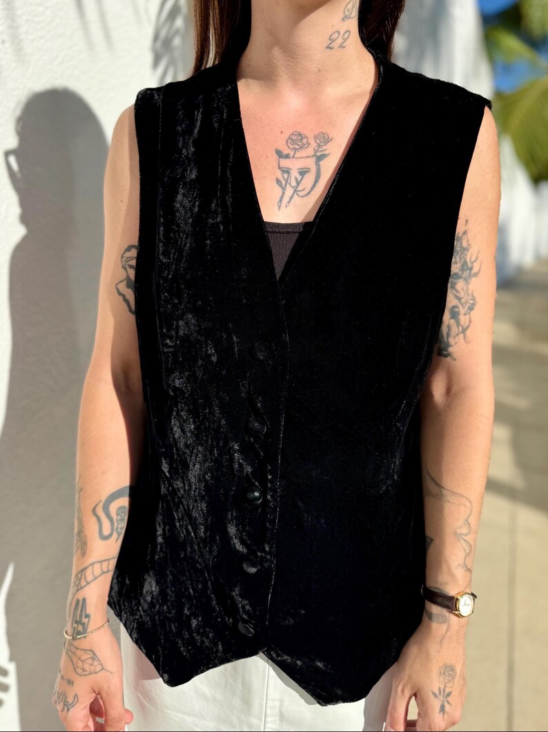 May include: A black velvet vest with a V-neck and button closure. The vest has a slightly textured appearance and is sleeveless. The garment is worn over a black top and white pants. The vest is a classic piece of apparel.
