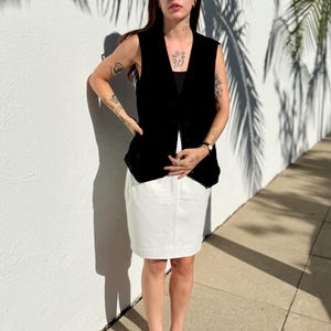 May include: A black sleeveless vest over a black top, paired with a white skirt. The outfit is completed with black sunglasses and flip-flops. The person is standing in front of a white wall with palm tree shadows.
