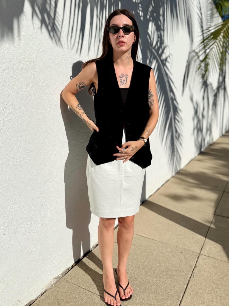 May include: A black sleeveless vest over a black top, paired with a white skirt. The outfit is completed with black sunglasses and flip-flops. The person is standing in front of a white wall with palm tree shadows.