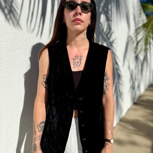 May include: A black velvet vest with a button-down front. The vest is sleeveless and has a V-neck. The person is wearing black sunglasses and a black top. The background is a white wall with shadows.