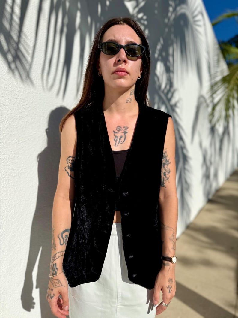 May include: A black velvet vest with a button-down front. The vest is sleeveless and has a V-neck. The person is wearing black sunglasses and a black top. The background is a white wall with shadows.