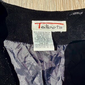 May include: Close-up of a black garment with a Talbots label. The label is white with red text and includes details about the fabric composition, which is 82% viscose rayon and 18% silk. The label also states the garment was made in the USA.