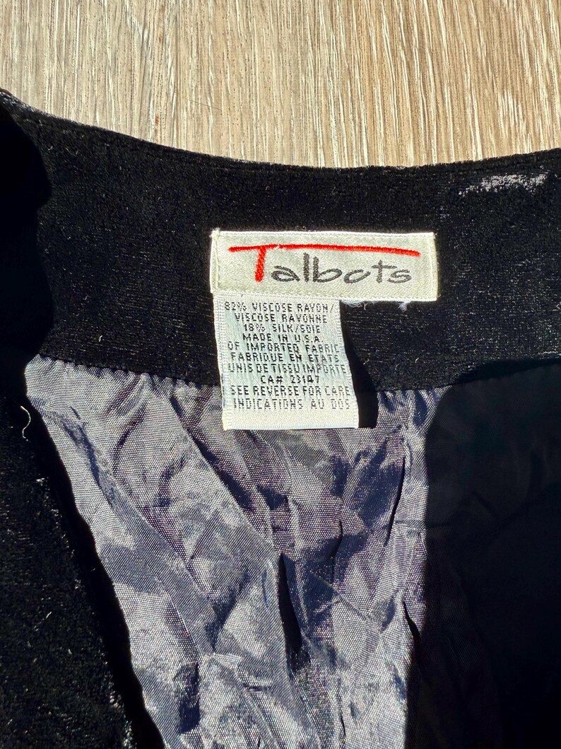 May include: Close-up of a black garment with a Talbots label. The label is white with red text and includes details about the fabric composition, which is 82% viscose rayon and 18% silk. The label also states the garment was made in the USA.