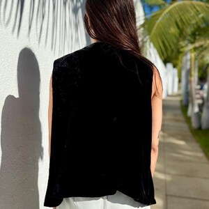 May include: A sleeveless black velvet top. The top has a draped design and a soft, luxurious texture. The person is wearing light gray pants. The background includes a white wall with shadows and palm trees.
