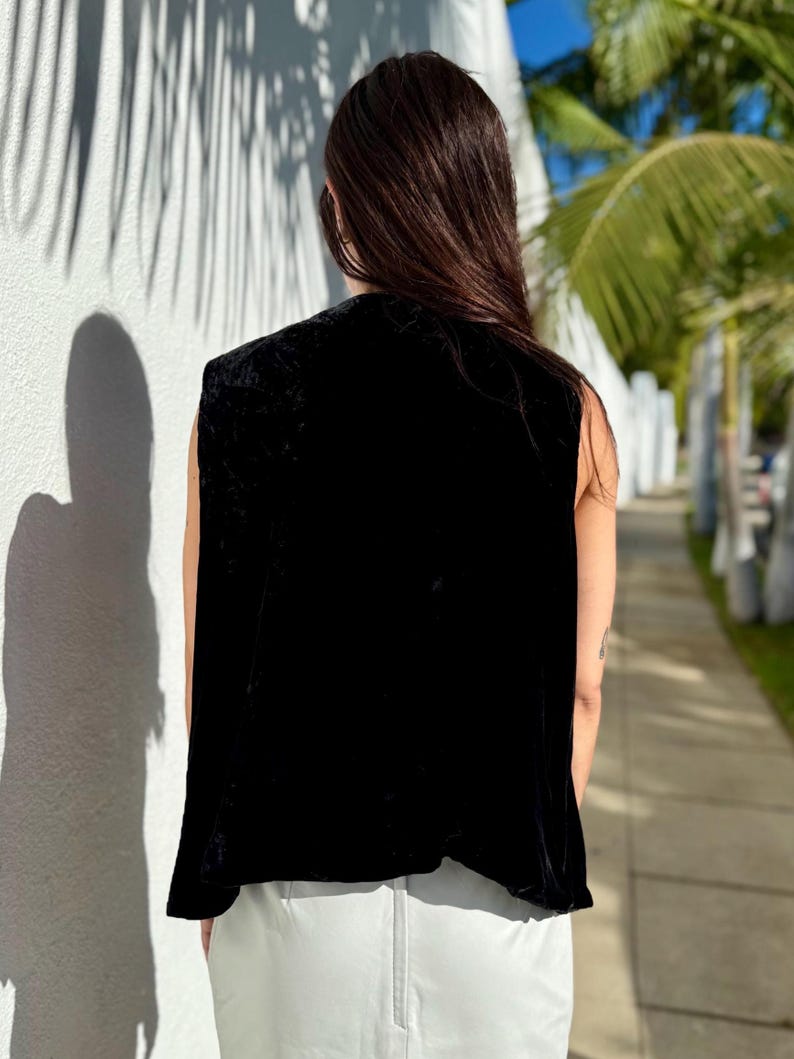 May include: A sleeveless black velvet top. The top has a draped design and a soft, luxurious texture. The person is wearing light gray pants. The background includes a white wall with shadows and palm trees.