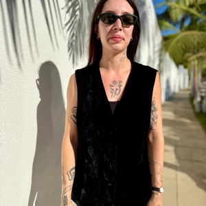 May include: A black velvet vest with a V-neck design. The vest is sleeveless and has a slightly textured appearance. The person is wearing sunglasses and white pants. The background includes a white wall and palm trees.