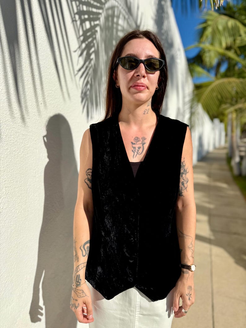 May include: A black velvet vest with a V-neck design. The vest is sleeveless and has a slightly textured appearance. The person is wearing sunglasses and white pants. The background includes a white wall and palm trees.