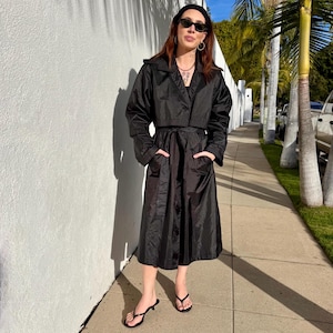 May include: A black trench coat with a cropped jacket top and a long skirt bottom. The coat has a belt, pockets, and a collar. The person is wearing black sunglasses, a black headband, and black sandals. The background is a white wall and a sidewalk.
