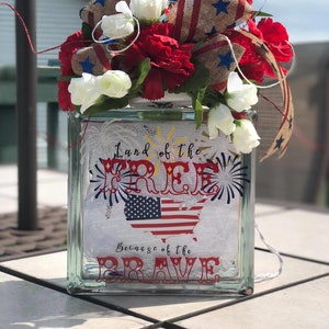 May include: A glass block vase with a patriotic theme. It features a red, white, and blue American flag design with the text "Land of the Free" and "Because of the Brave". The vase is decorated with red, white, and blue carnations and a burlap bow with stars.