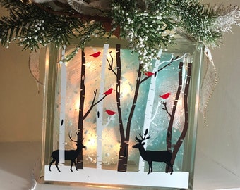 May include: A glass block with a winter scene painted on it. The scene includes a snowy forest with birch trees, red cardinals, and two deer. The block is lit from within, creating a warm glow. The block is decorated with a white and gold ribbon and a snowflake.