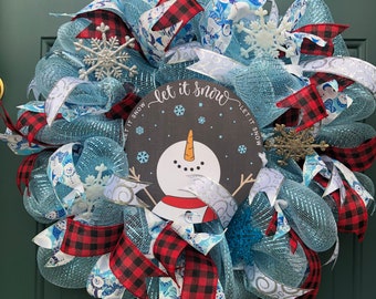 May include: A winter wreath with a snowman design. The wreath features blue mesh, red and black plaid ribbons, and snowflake accents. A circular sign in the centre reads "Let it snow" and depicts a snowman.