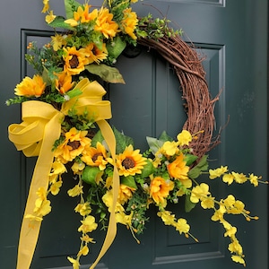 May include: A sunflower wreath with a yellow ribbon bow and yellow flower accents. The wreath is made of grapevine and is decorated with artificial sunflowers and yellow flowers.