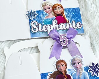 May include: Two white favor boxes with a Frozen theme. Each box has a picture of Elsa and Anna from the movie Frozen. The boxes are decorated with purple bows, glitter, and the name "Stephanie" and the number "5".
