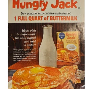 May include: A vintage advertisement for Hungry Jack Buttermilk Pancake Mix. The ad features a stack of golden brown pancakes with a pat of butter on top, a bottle of buttermilk, and a box of Hungry Jack pancake mix. The text on the ad reads "New from Pillsbury - Hungry Jack. New pancake mix contains equivalent of 1 FULL QUART of BUTTERMILK. It's so rich in buttermilk the only liquid you add is water!"