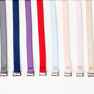 Może przedstawiać: Kolekcja ramiączek od biustonoszy w różnych kolorach, w tym czarnym, szarym, granatowym, fioletowym, czerwonym, jasnoniebieskim, różowym, beżowym i białym. Każde ramiączko ma metalowe zapięcie. Ramiączka ułożone są pionowo na białym tle.