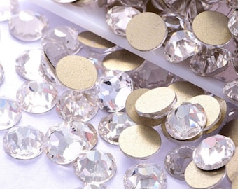 May include: A close-up of a large quantity of clear glass rhinestones, some with a gold backing. The rhinestones are scattered on a white surface.