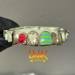 May include: Custom gold grillz with unique designs. One tooth features a red smiley face, another a heart, and a third with blue and green stripes. The brand name "UNIK GRILLZ" is displayed in a yellow and orange font.