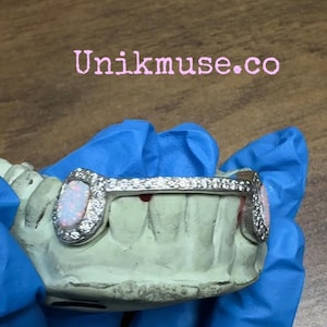 May include: Silver-colored grill with opal-like stones and clear crystals. The grill is set on a light green dental mold. The text "Unikmuse.co" is displayed in pink above the grill.